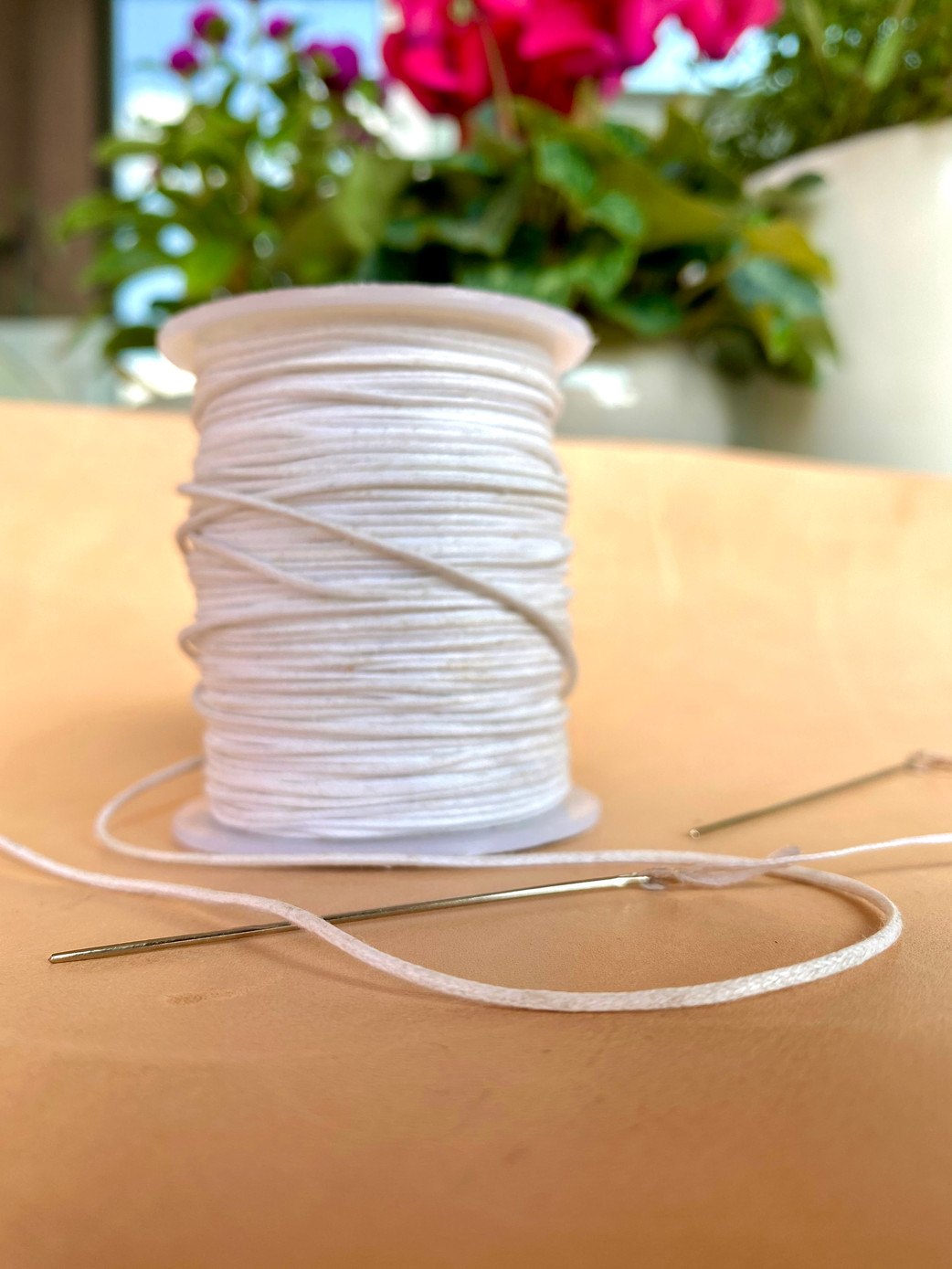 A spool of white braided thread on vegetable-tanned leather, with two threaded needles in front of it
