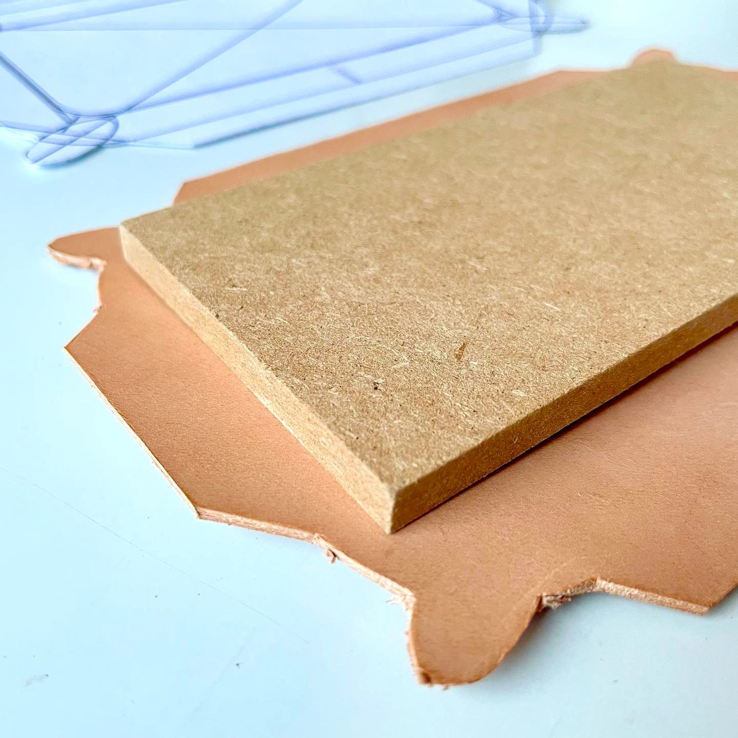 A piece of vegetable-tanned leather with its edges still uncut, under a block of fibreboard on a white table, with paper templates behind it