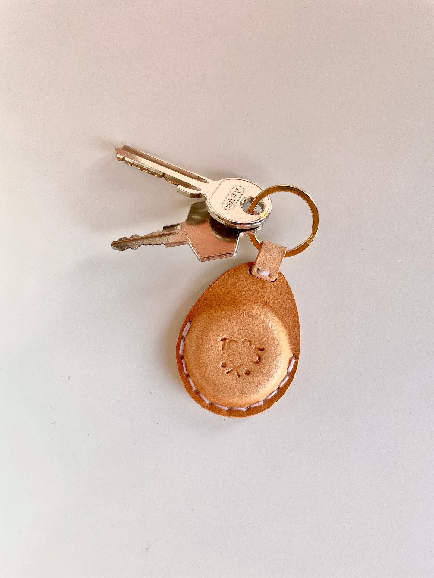 AirTag key fob by 1985 Studio from above, the 1985 mark stamped into its vegetable-tanned leather, two keys on its brass ring
