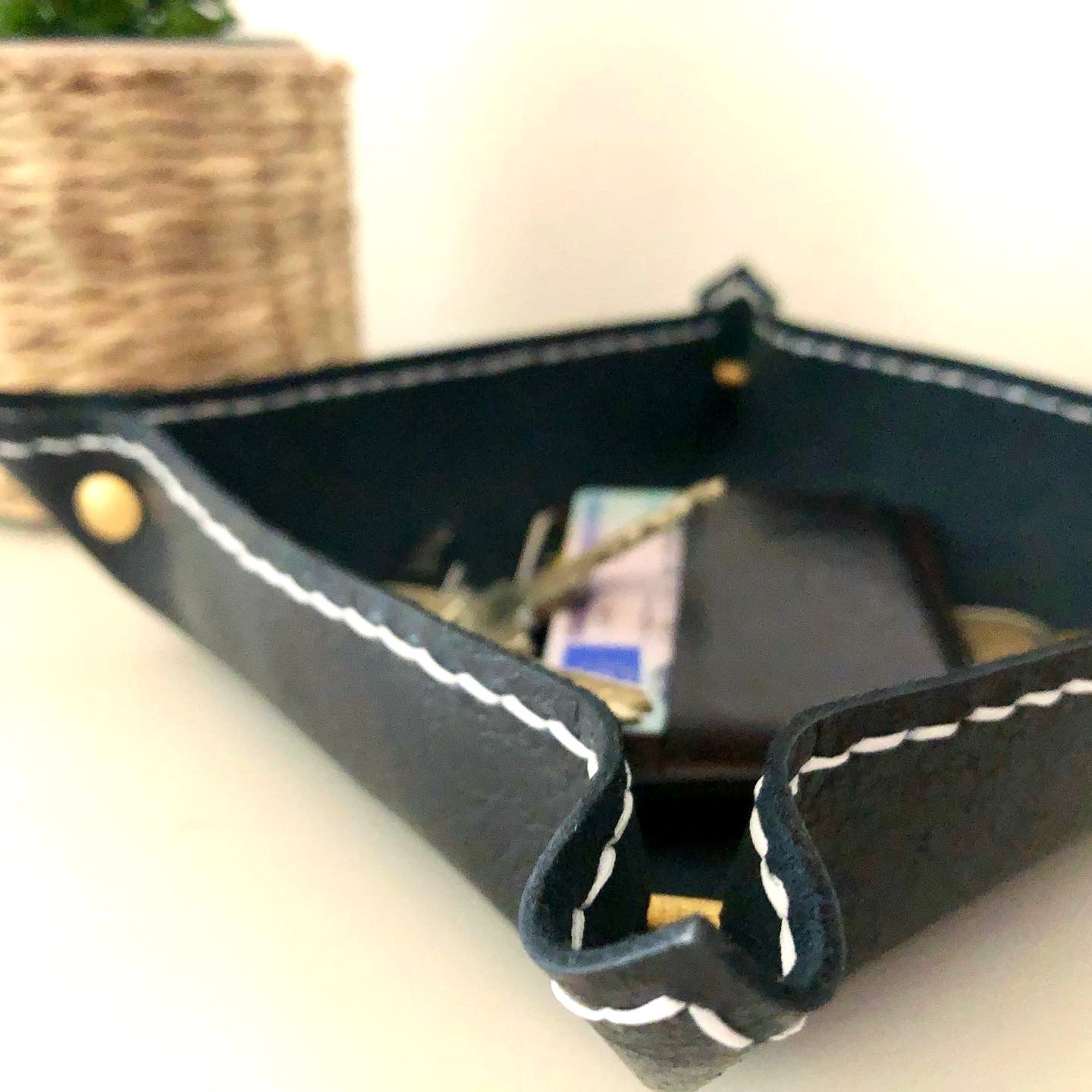 Black valet tray by 1985 Studio at table height, chrome-tanned leather with a brass Chicago screw at each raised corner and white stitching along its edge, keys and a card wallet inside