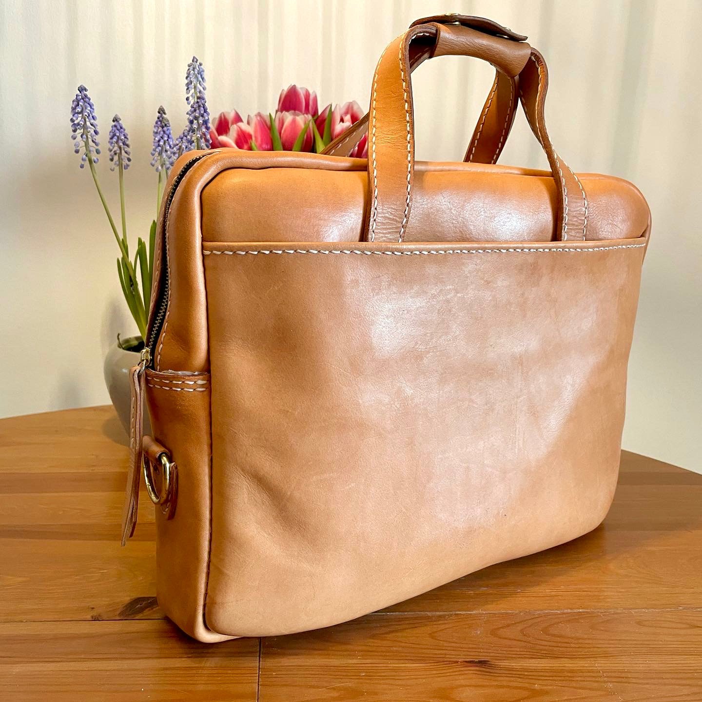 Munich Bag by 1985 Studio from the back at an angle, vegetable-tanned leather with its zip running along the top and a ring for the strap at the side, tulips and grape hyacinths behind it