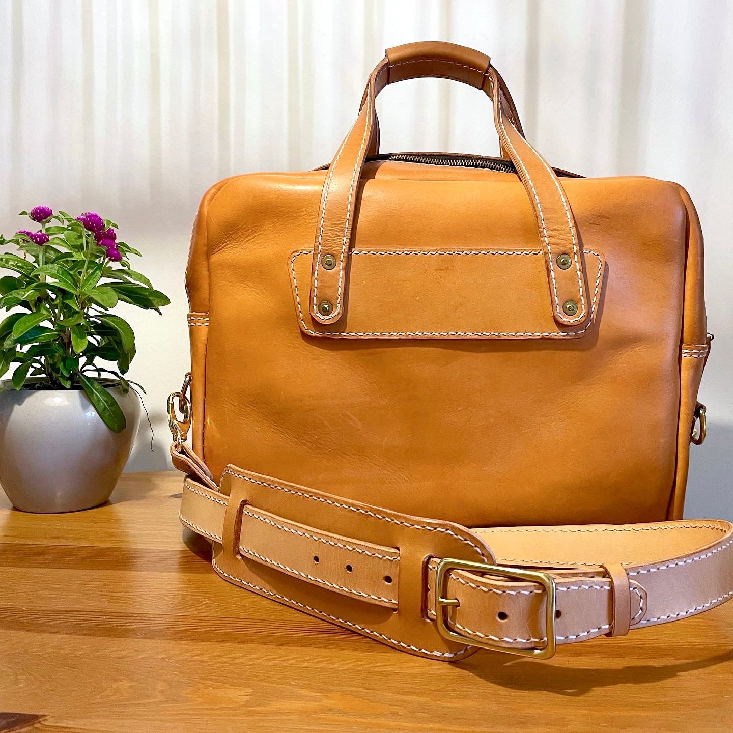Munich Bag by 1985 Studio from the front, a panel of vegetable-tanned leather riveted on below the handles and its shoulder strap with a brass buckle lying in front