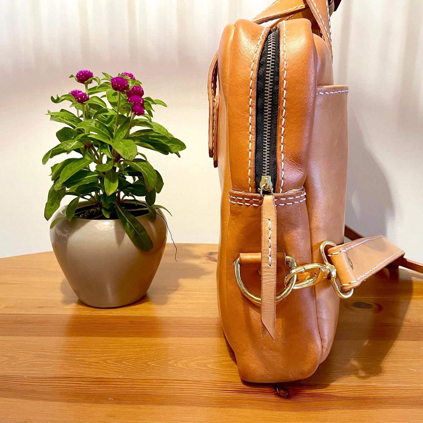 Munich Bag by 1985 Studio in vegetable-tanned leather from the side: the zip down its end, a brass ring and snap hook for the strap, and a potted plant beside it