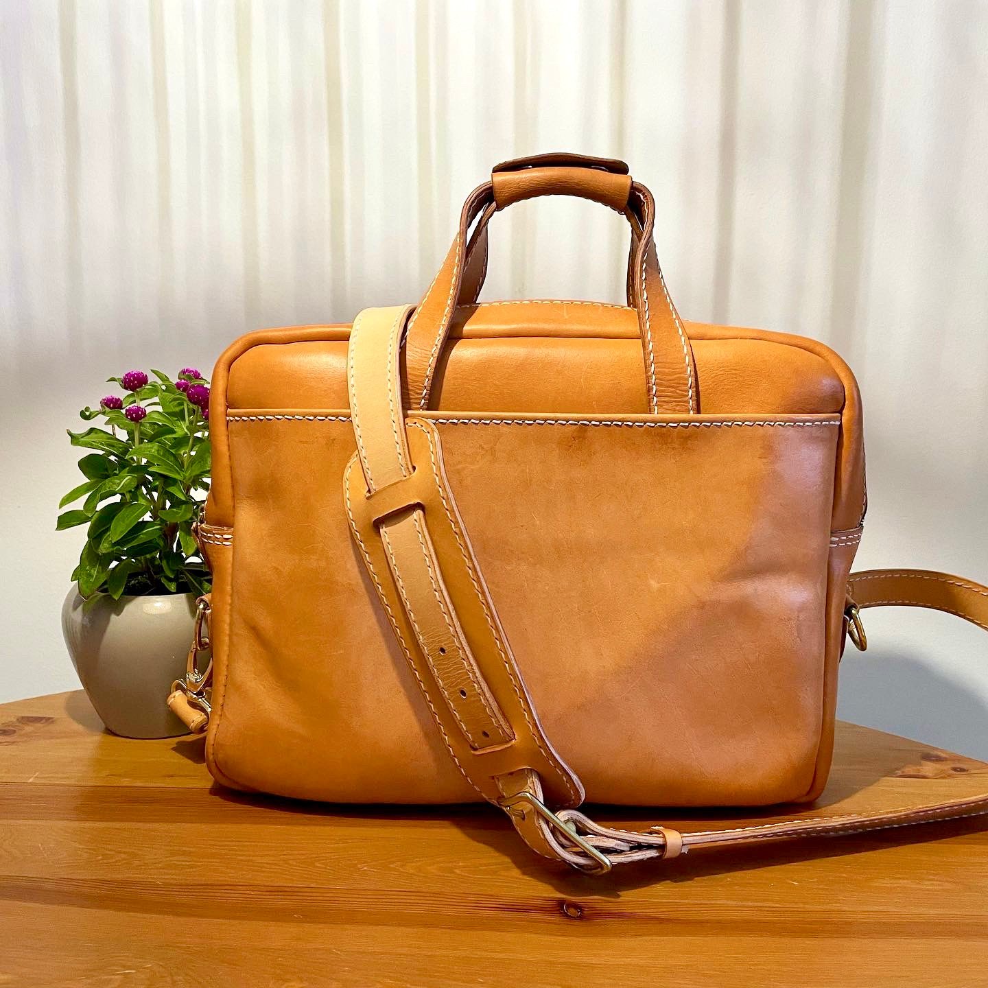 Munich Bag by 1985 Studio in vegetable-tanned leather from the front: two top handles, a shoulder strap across it and a pot of purple flowers beside it on a wooden table