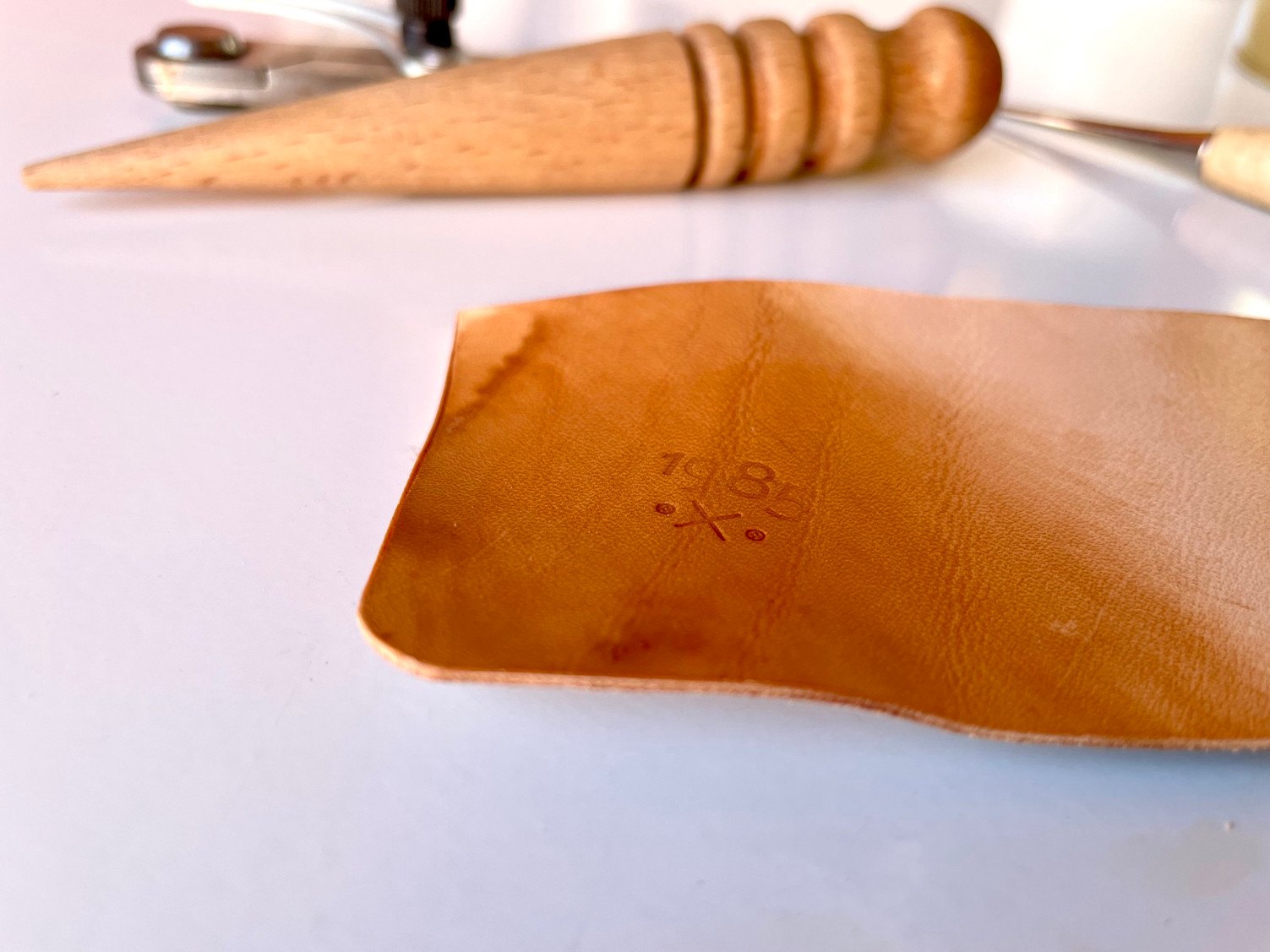 A piece of vegetable-tanned leather darkened with oil and beeswax and stamped with the 1985 Studio mark, in front of a wooden edge slicker