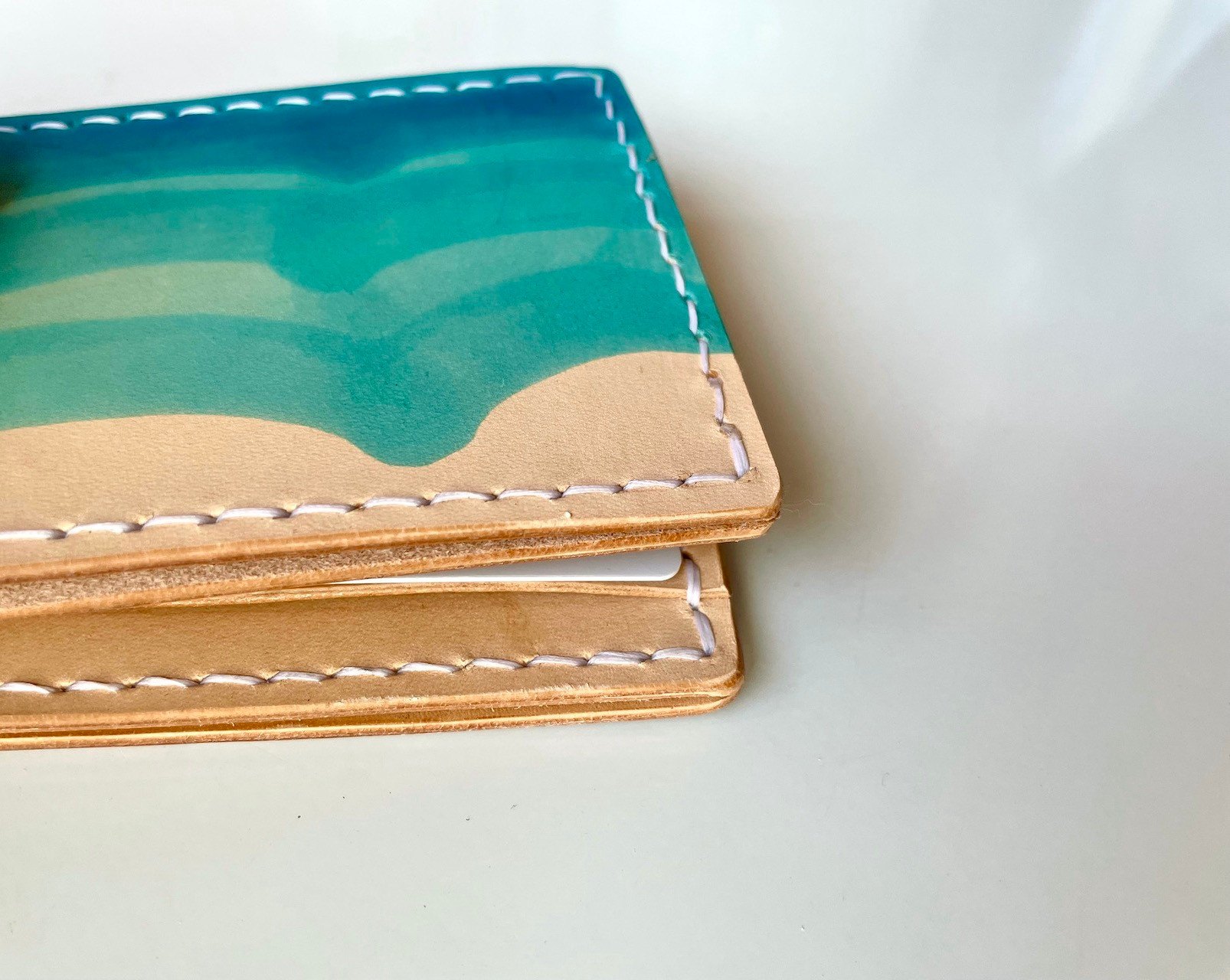 A corner of the Wave Wallet by 1985 Studio up close, turquoise waves dyed into the vegetable-tanned leather above its natural color, the edge stitched in white