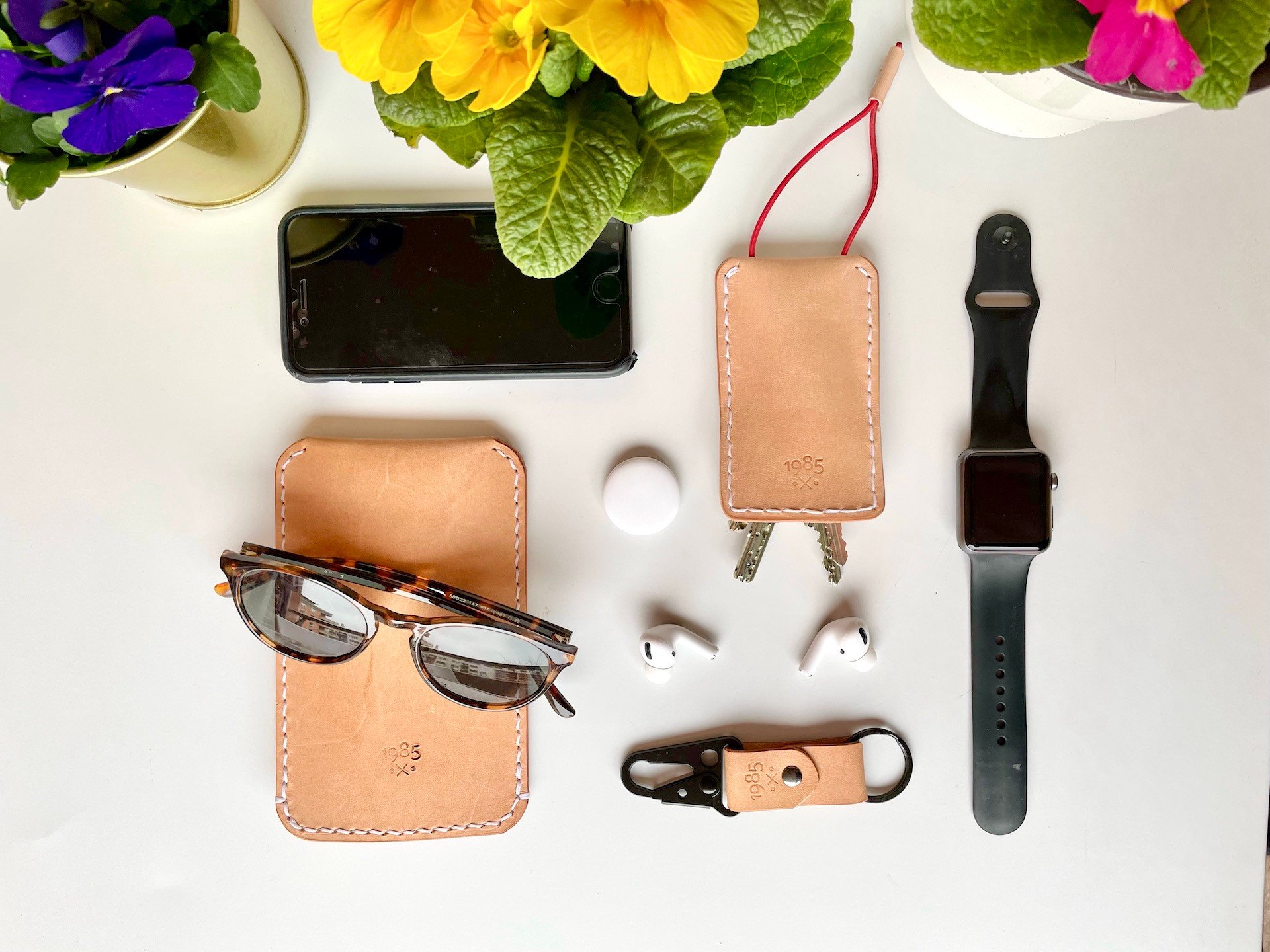 Handmade leather goods by 1985 Studio on a white table under spring flowers: a sunglasses pouch under tortoiseshell sunglasses, a key case with a red pull cord and two keys, and a key clip with a black carabiner, beside an iPhone, an AirTag, AirPods and an Apple Watch