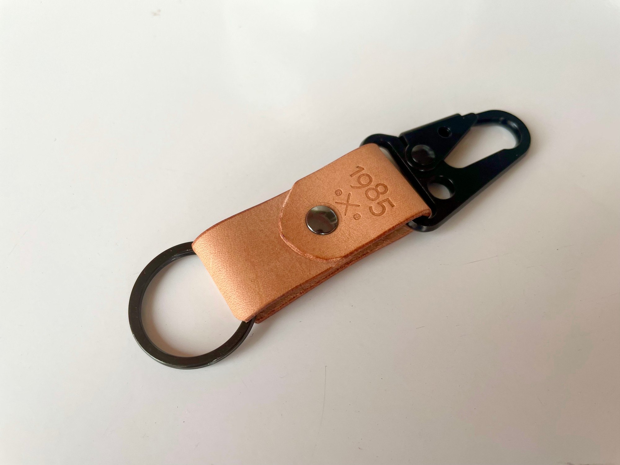 Carabiner keychain by 1985 Studio up close: the black clip, the riveted strap of vegetable-tanned leather with the 1985 mark, and a black split ring