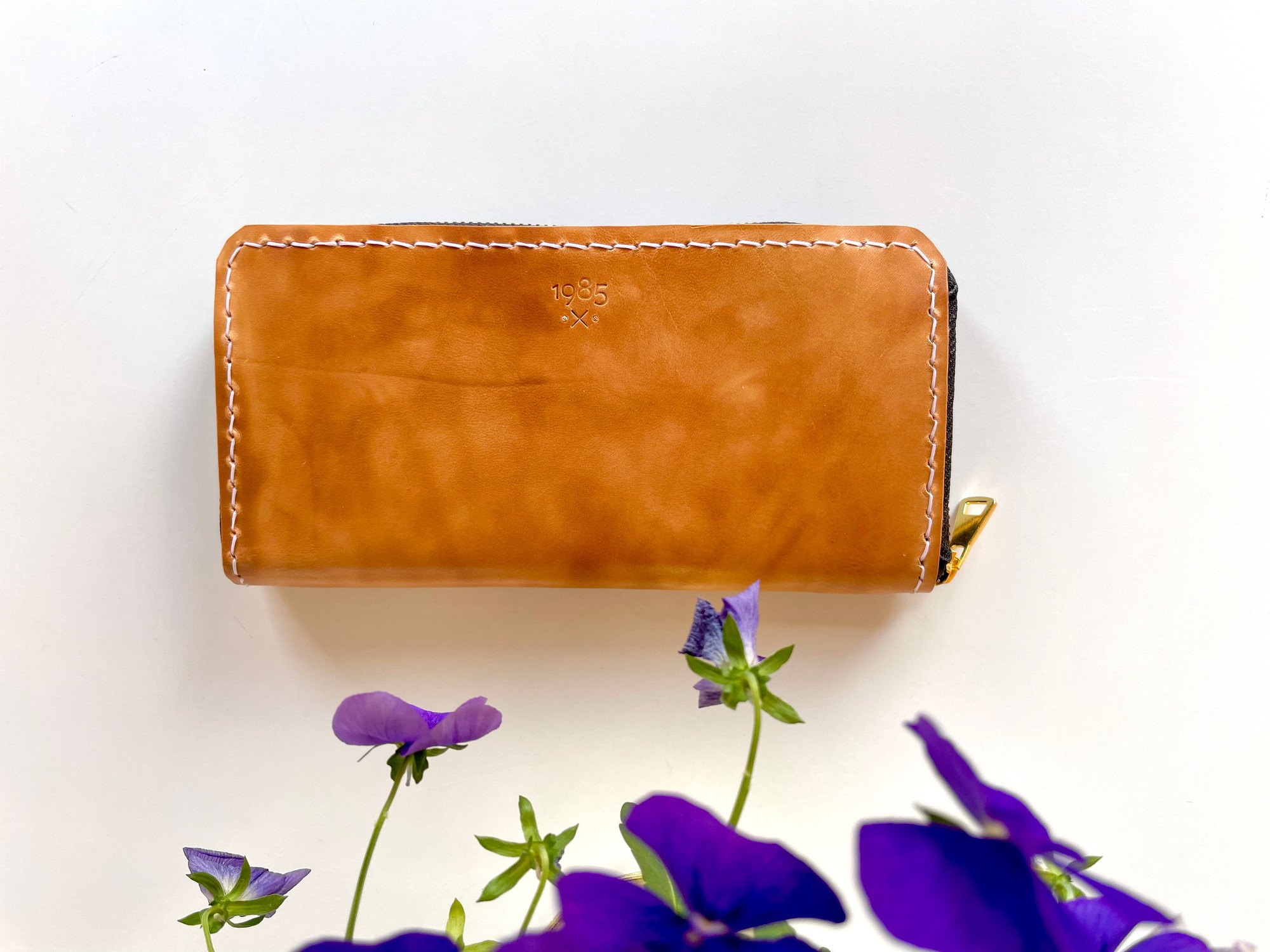 Zip-around long wallet by 1985 Studio in vegetable-tanned leather, closed on a white table, purple pansies in front of it