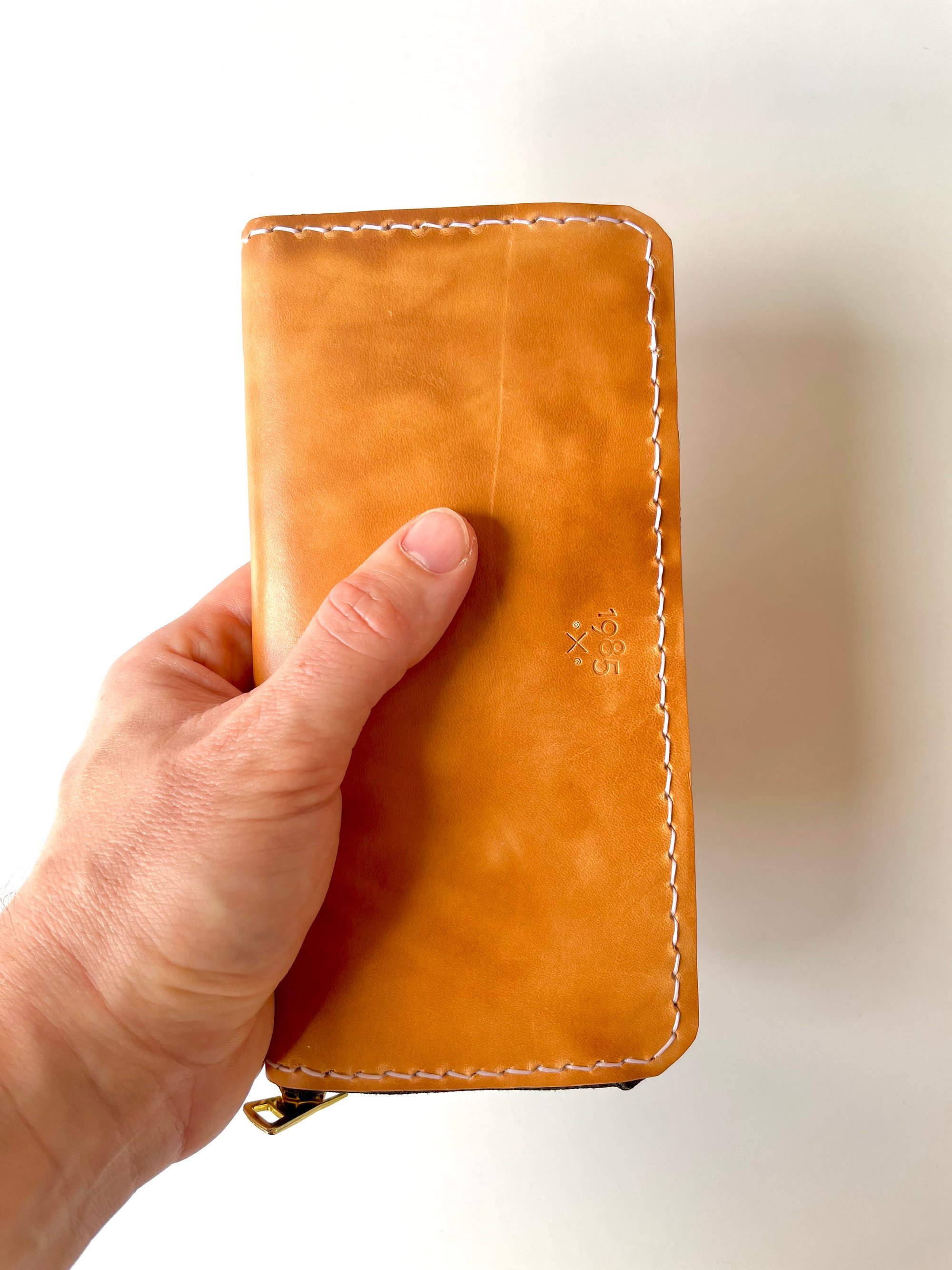 Zip-around long wallet by 1985 Studio in vegetable-tanned leather held in a hand, its mottled cognac shell of hot-stuffed leather stamped with the 1985 mark and stitched in pale thread