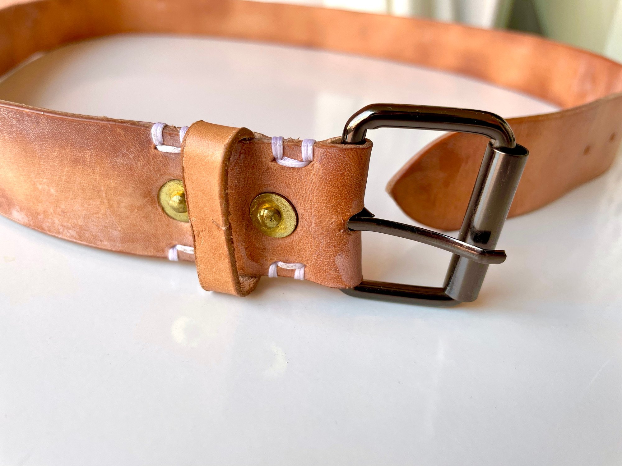 The gunmetal roller buckle of a belt by 1985 Studio up close, the vegetable-tanned leather folded around it and held by two brass rivets beside white stitches