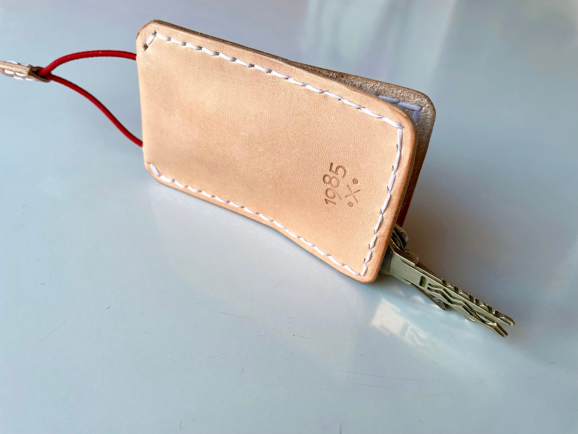 Slim key case by 1985 Studio in vegetable-tanned leather on a white table, a key reaching out of the bottom and the red cord out of the top