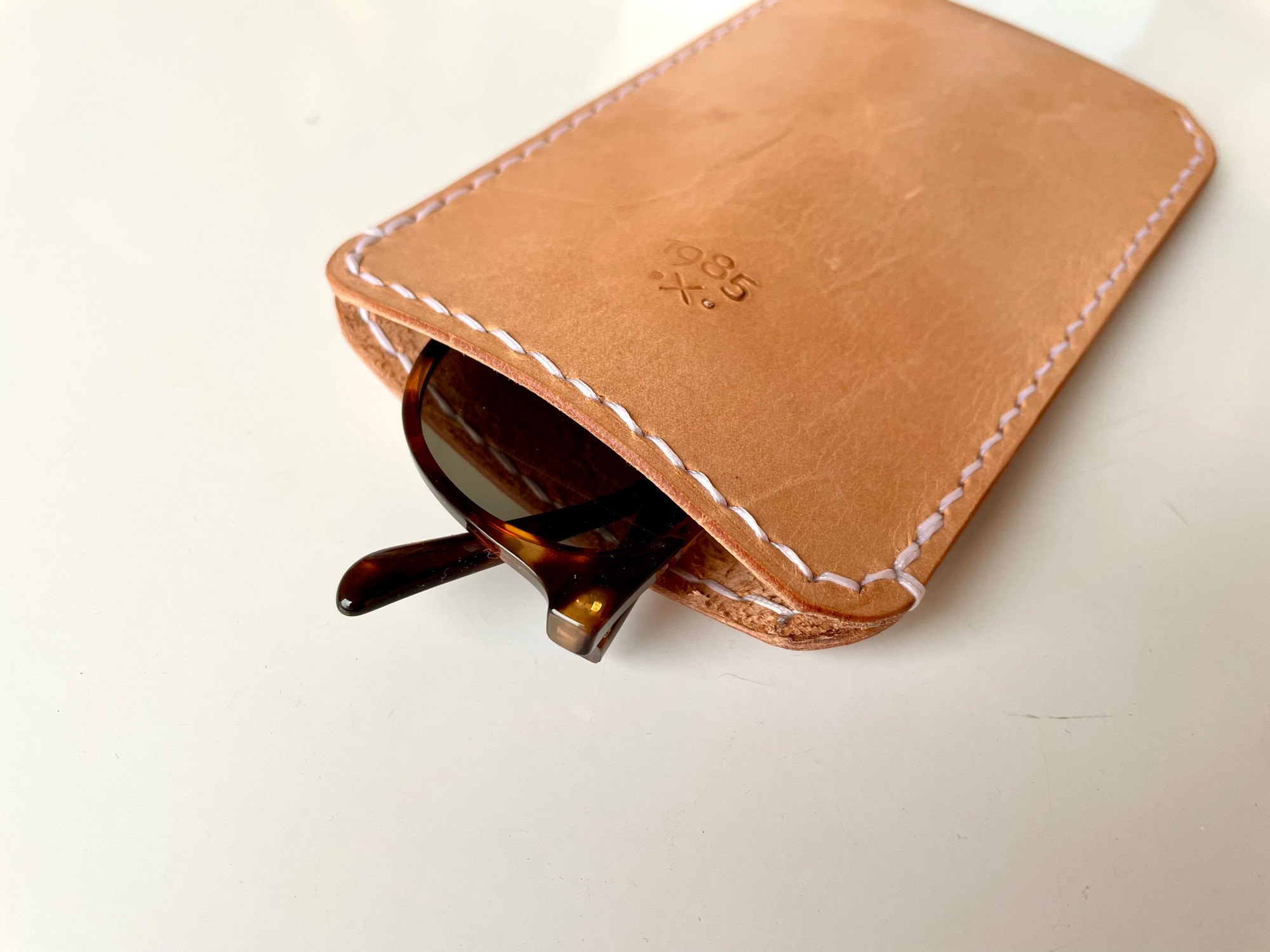 Slim sunglasses pouch by 1985 Studio, sunglasses sliding out of its opening, the 1985 mark stamped into the vegetable-tanned leather
