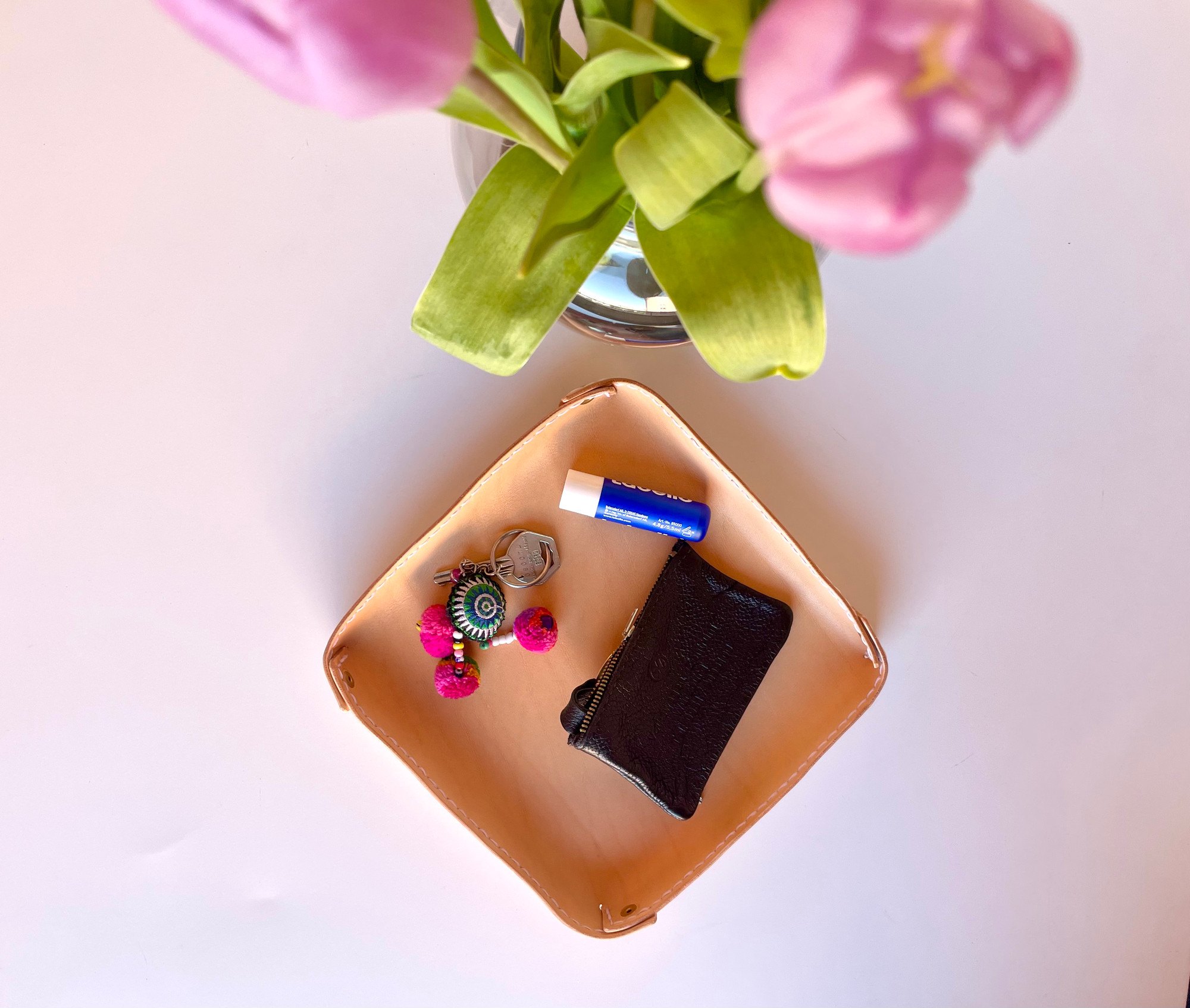 Fold-over valet tray by 1985 Studio from above, a square of vegetable-tanned leather with its corners folded up and a few pocket things inside, below pink tulips