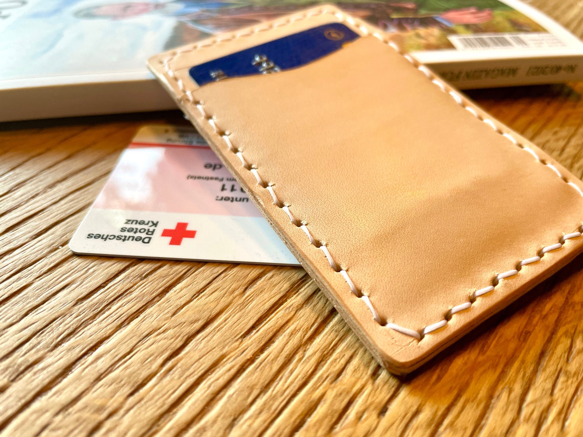 Vertical card wallet by 1985 Studio on a wooden table, a card in its front slot of natural vegetable-tanned leather, white stitching along the edge
