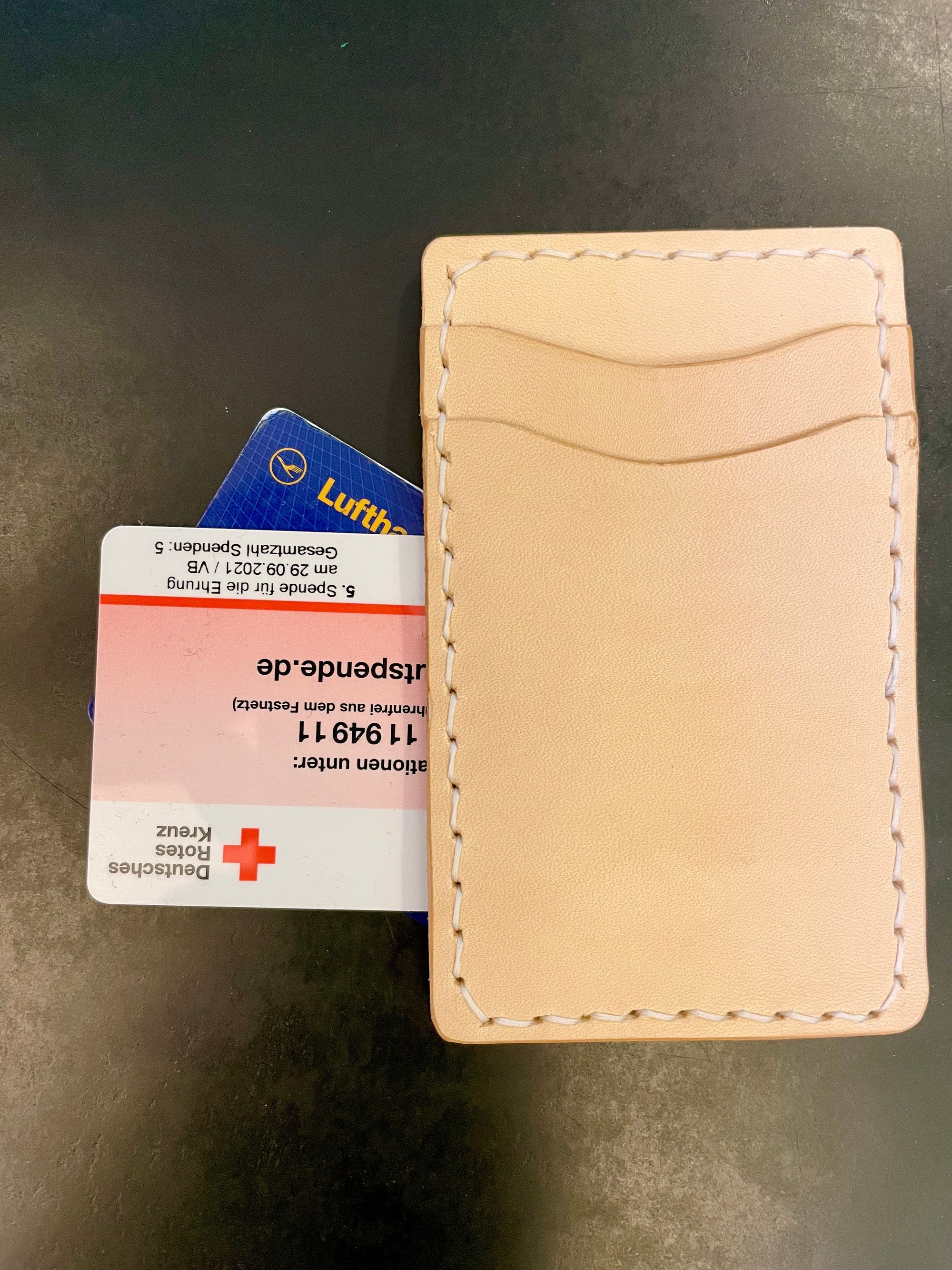 Vertical card wallet by 1985 Studio on a dark table, natural vegetable-tanned leather stitched in white, two curved slots on its back and cards behind it