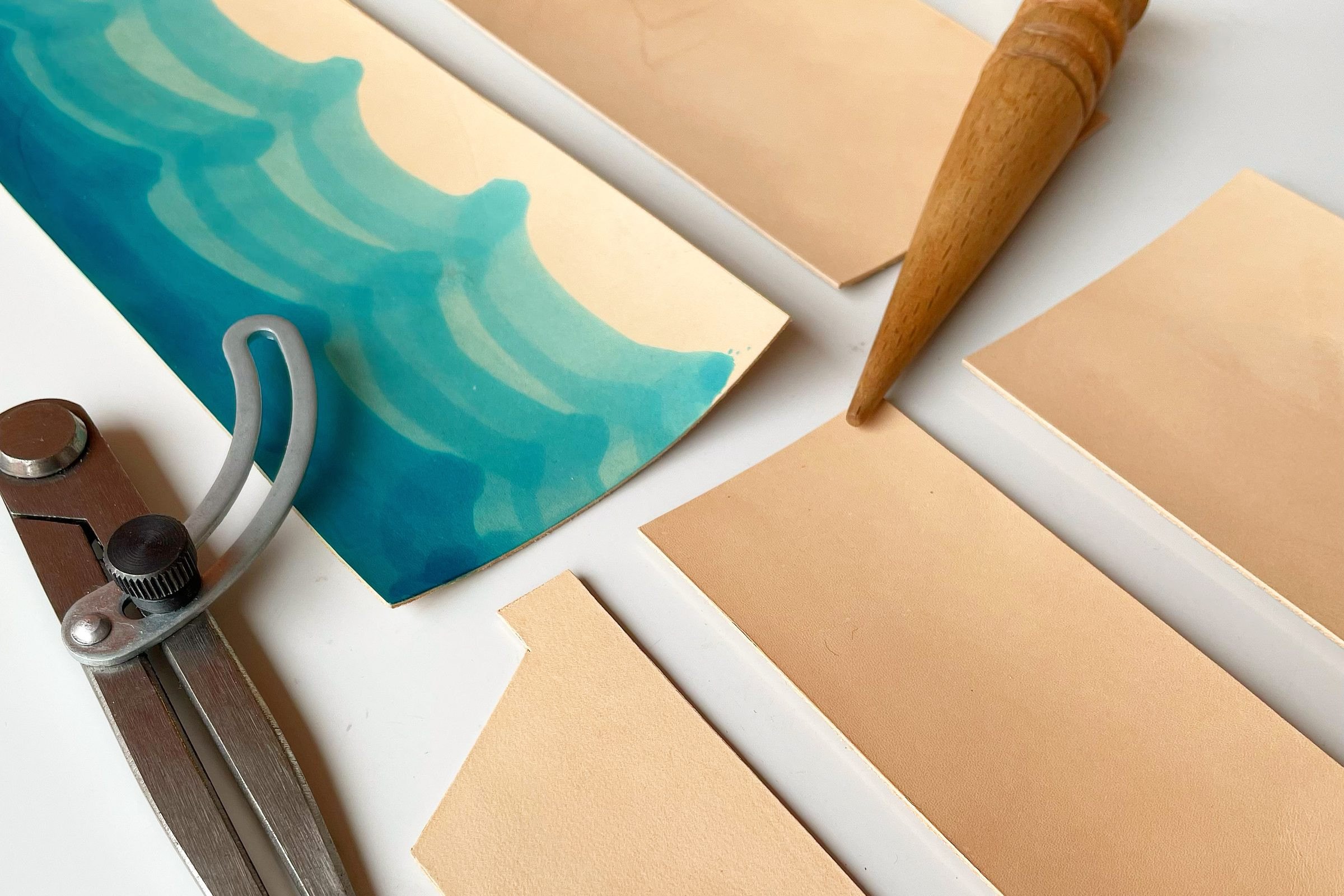 Pieces of vegetable-tanned leather at 1985 Studio, one dyed in teal waves, on a white bench beside the curved arm of a wing divider