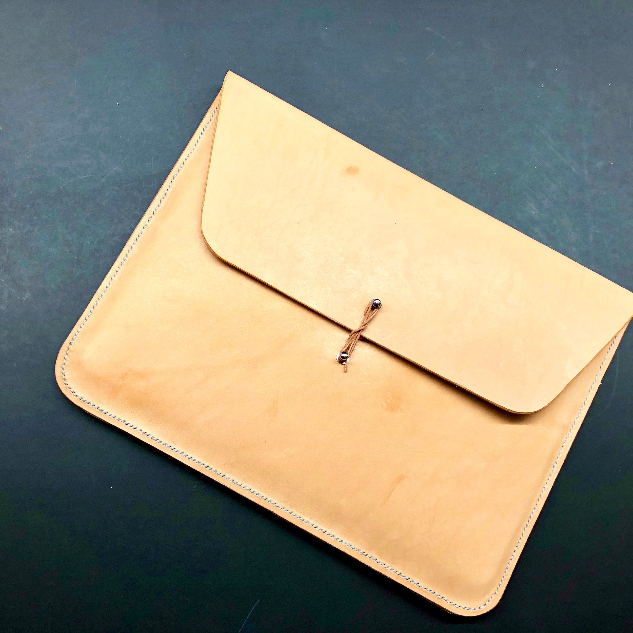 Envelope laptop case by 1985 Studio from the front on a dark surface, natural vegetable-tanned leather with its flap closed by a string wound around two metal studs