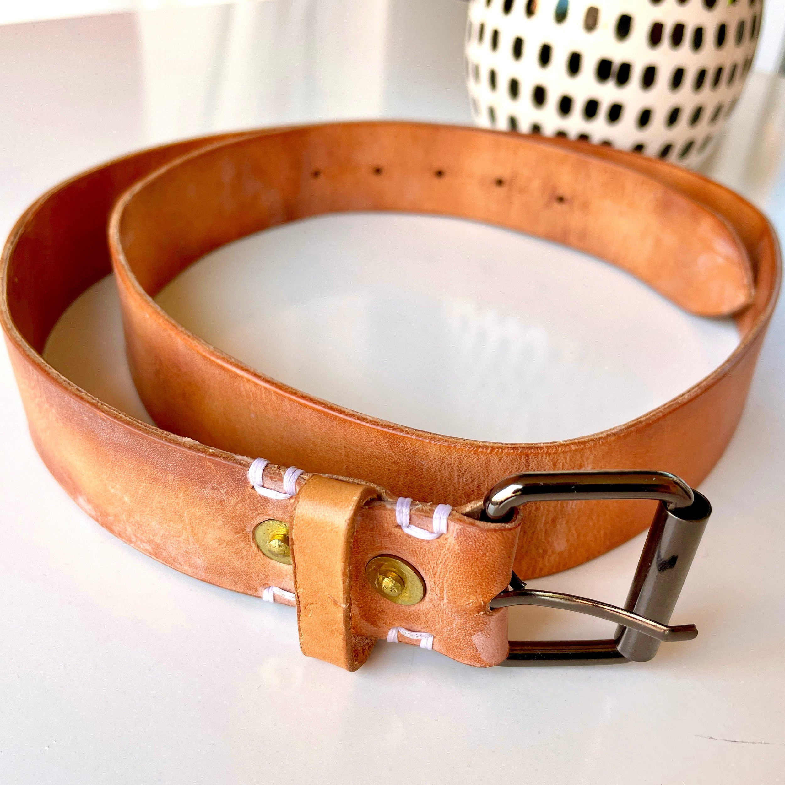 Roller buckle belt by 1985 Studio coiled on a white table, thick vegetable-tanned leather with a gunmetal roller buckle, two brass rivets and white stitches at the fold