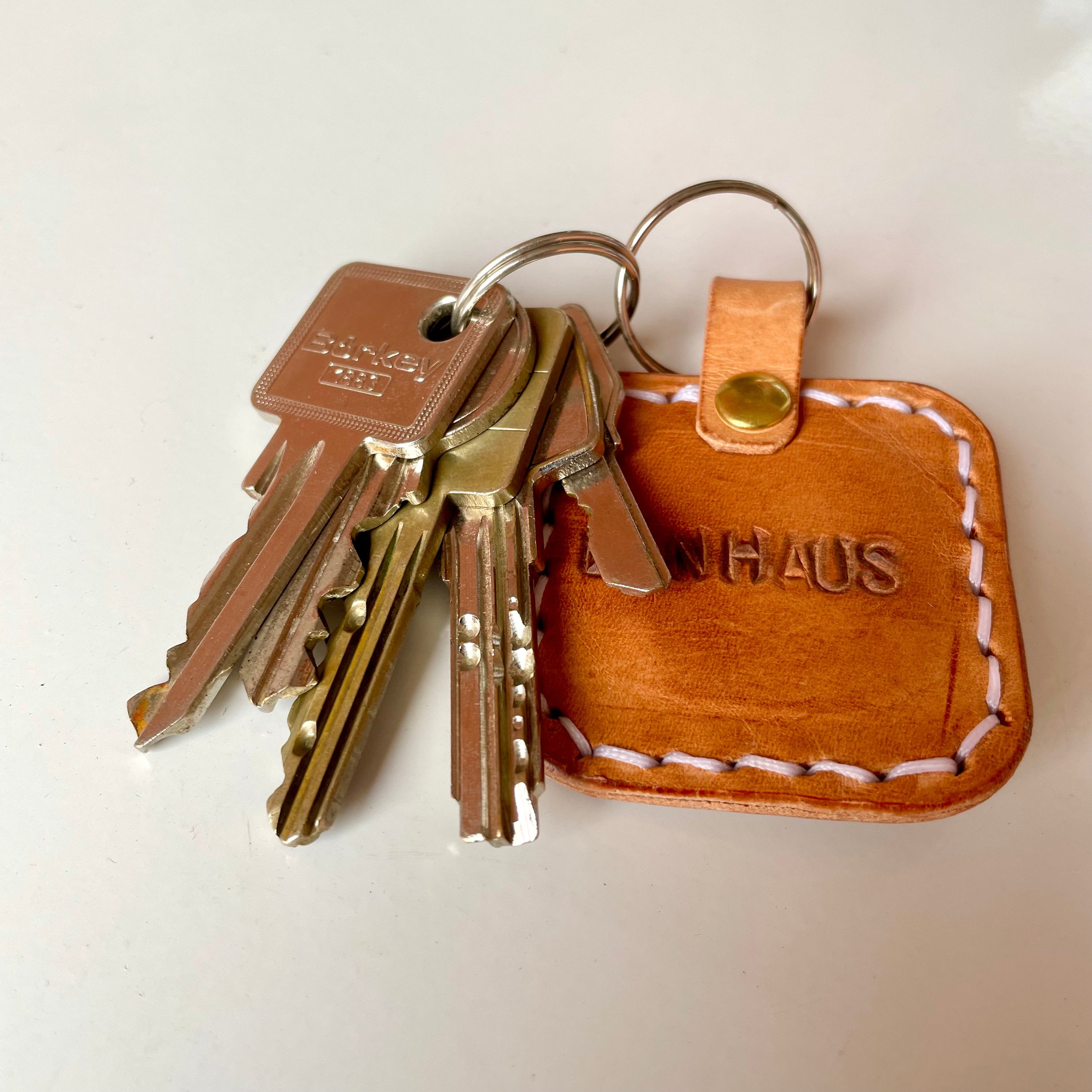 Square key tag by 1985 Studio in cognac vegetable-tanned leather, stitched in white around a stamped word, its loop held by a brass rivet, next to a bunch of keys