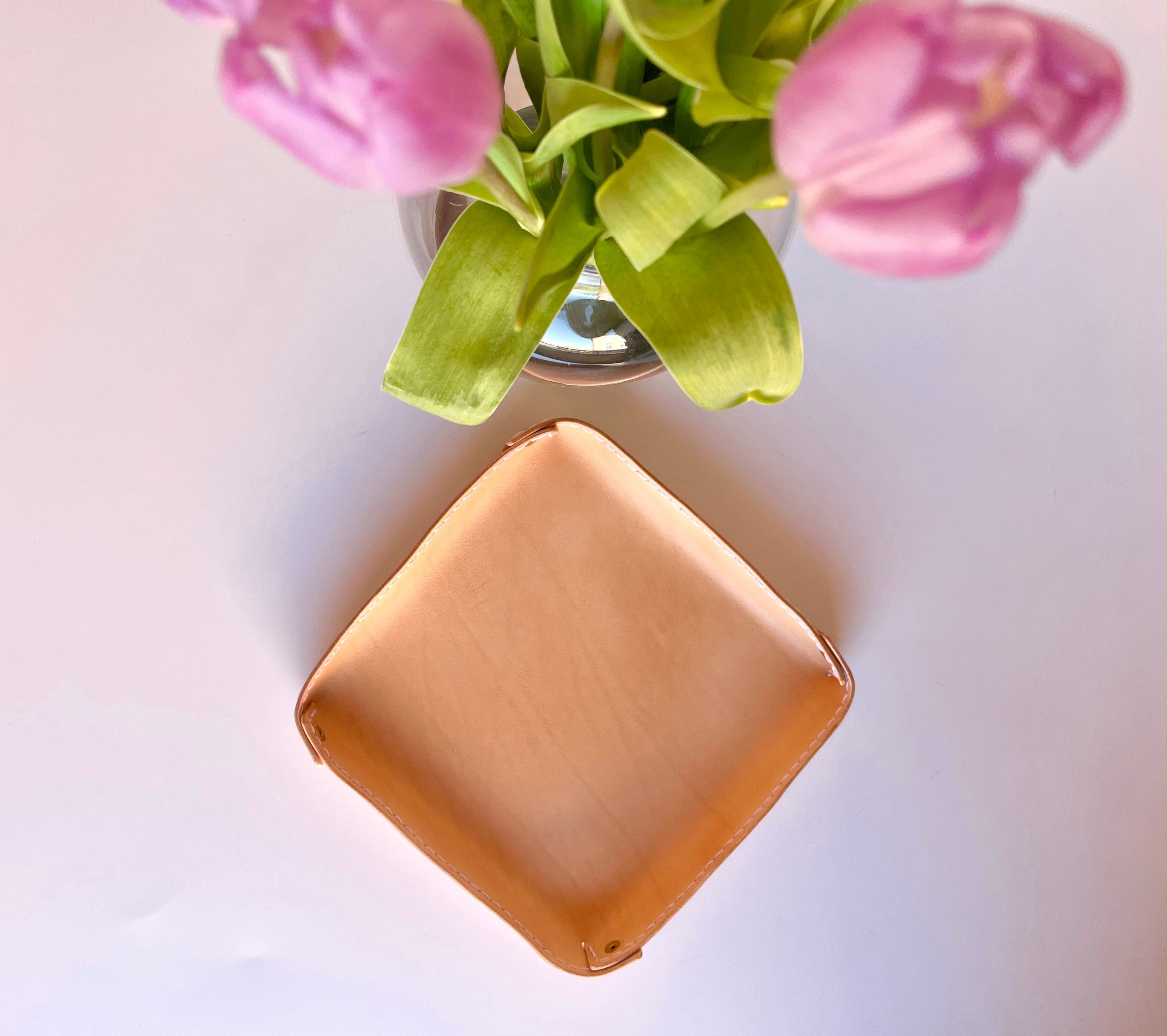 Square fold-over valet tray by 1985 Studio in vegetable-tanned leather, its corners held up by brass rivets, below a vase of pink tulips