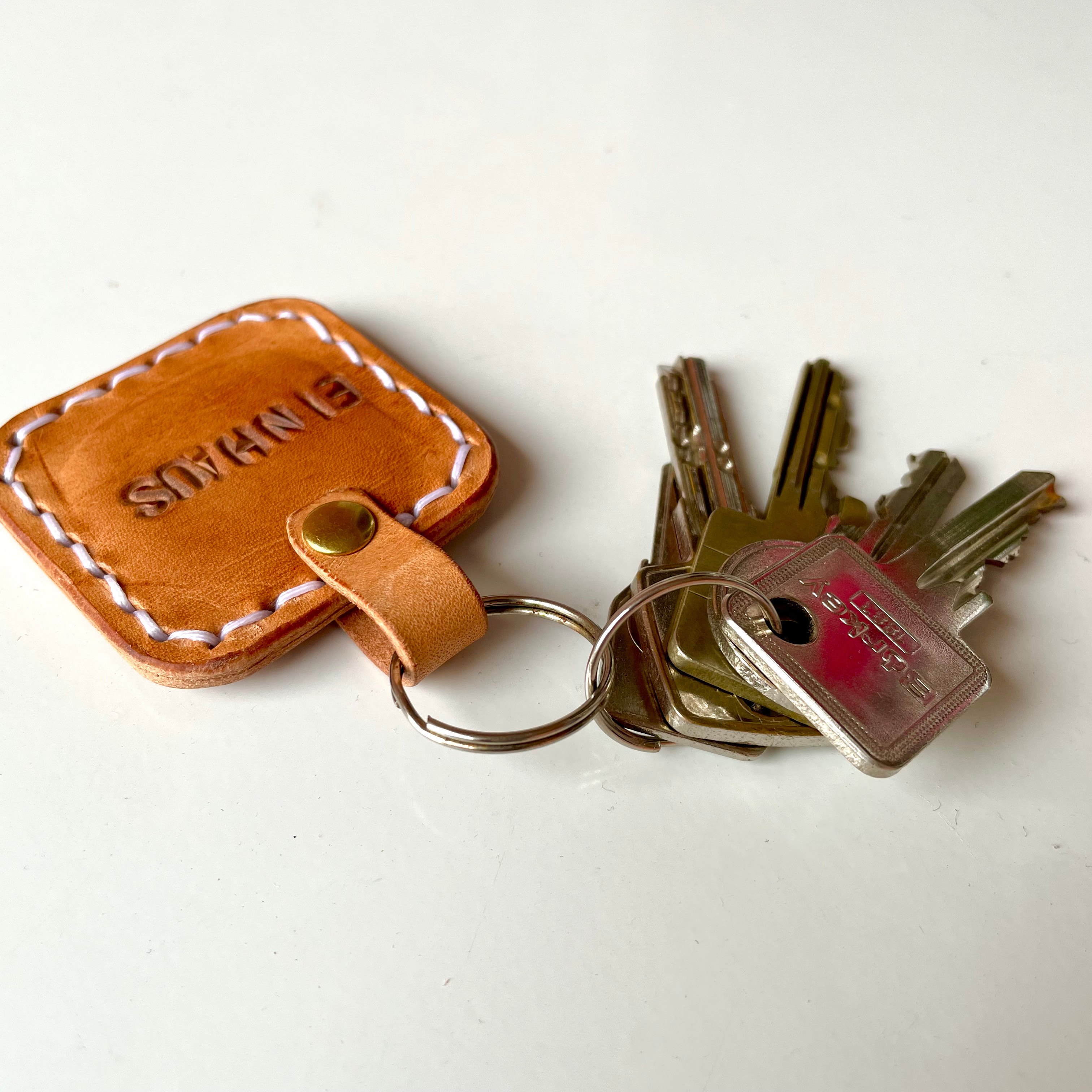 Square key tag by 1985 Studio in vegetable-tanned leather, upside down beside its keys, the stamped word in a frame of white stitches and the loop held by a brass rivet