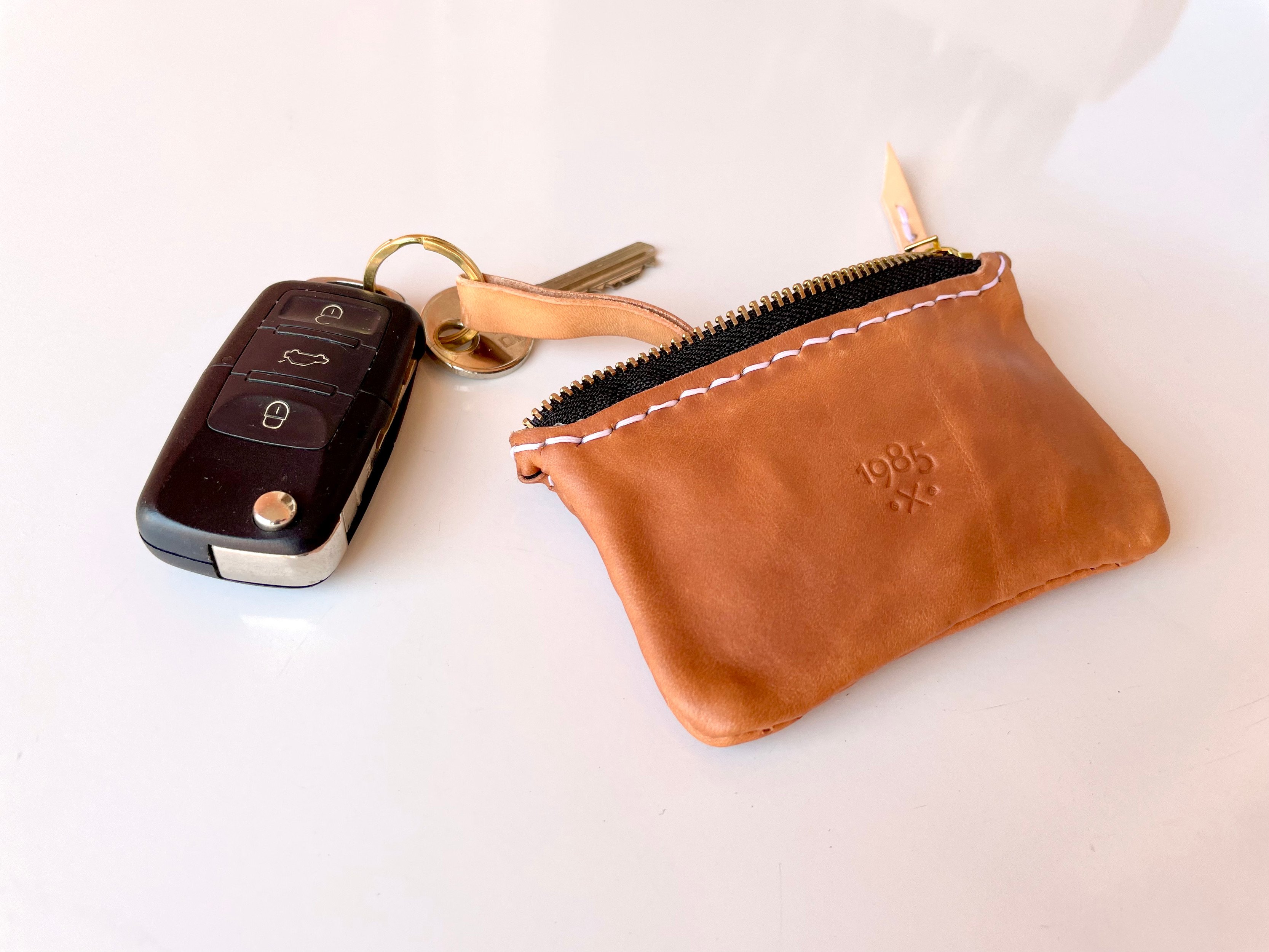 Car key pouch by 1985 Studio in cognac vegetable-tanned leather stamped with the 1985 mark, its zip open, a car key and a house key hanging out on a brass ring