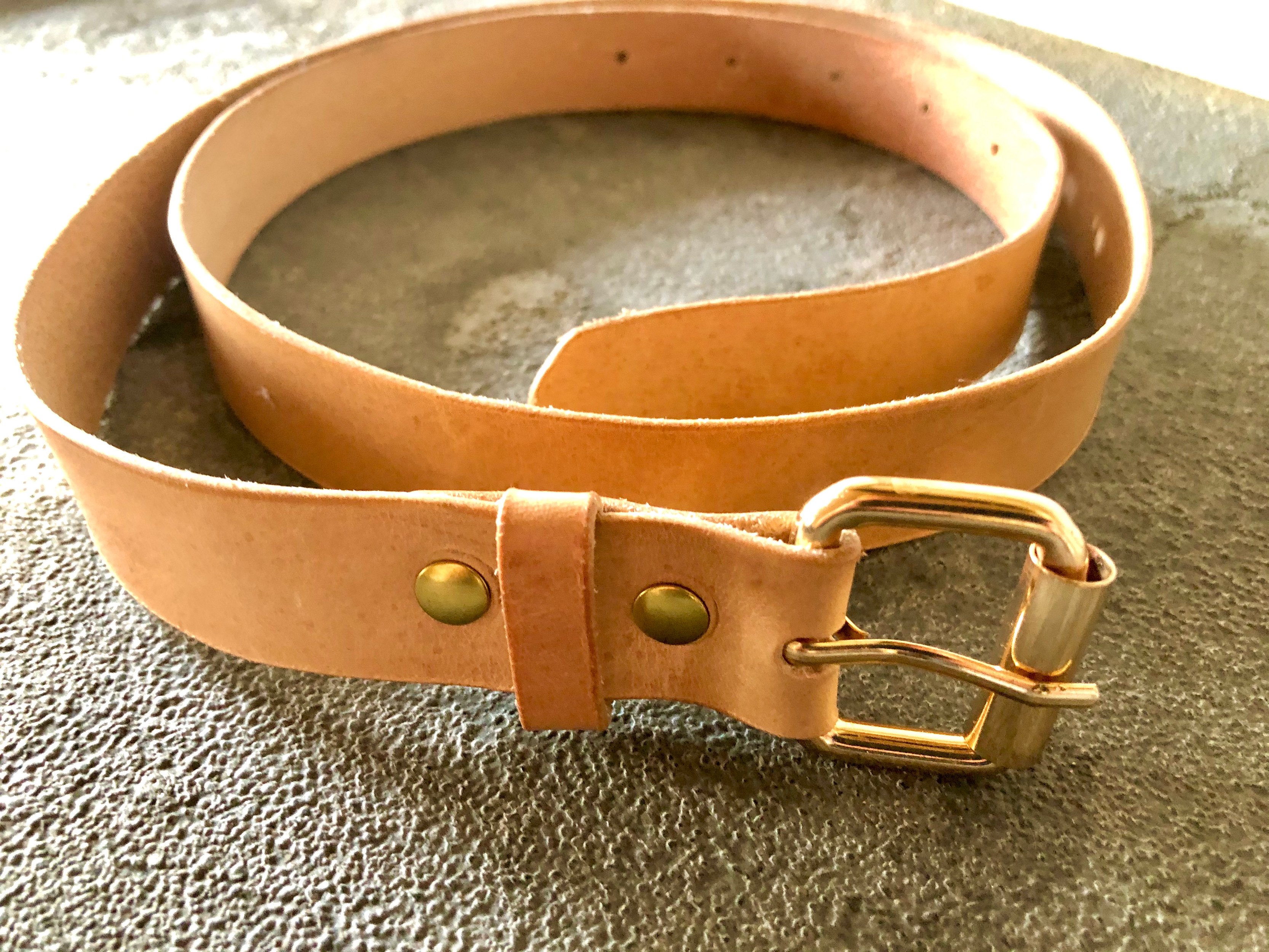 Natural leather belt by 1985 Studio coiled on grey stone, pale vegetable-tanned leather with a brass roller buckle, a keeper and two brass rivets