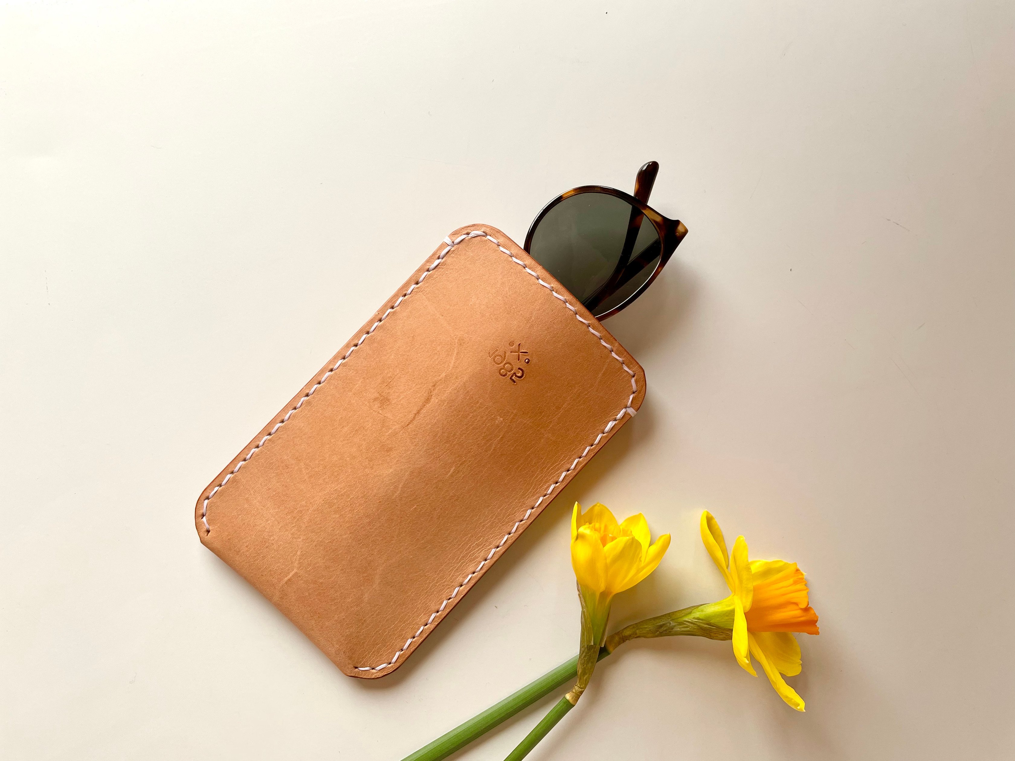 Slim sunglasses pouch by 1985 Studio from above, vegetable-tanned leather saddle-stitched by hand, sunglasses half out of it beside yellow daffodils
